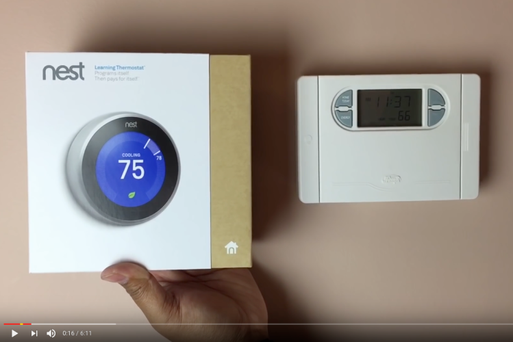 Screen capture from installation video on YouTube: a hand holding up a Nest thermostat, still in its box, near the thermostat to be replaced