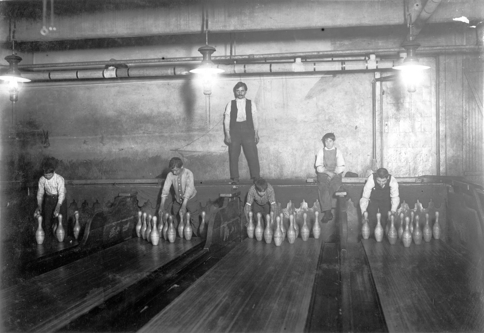 Four teenage boys setting pins by hand in a New York City bowling alley, 1910, under the supervision of a boss